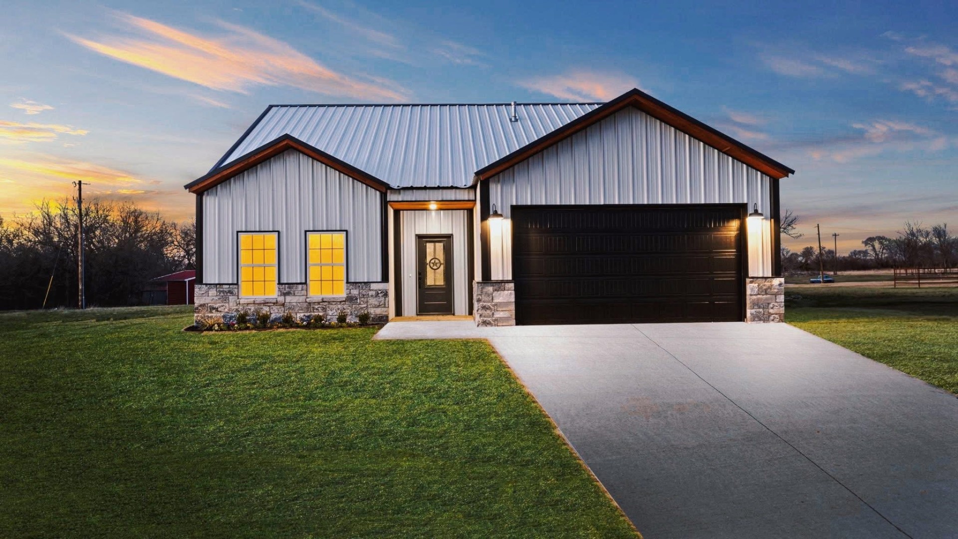 Modern barndominium with white metal siding, stone wainscot and black garage door at sunset
